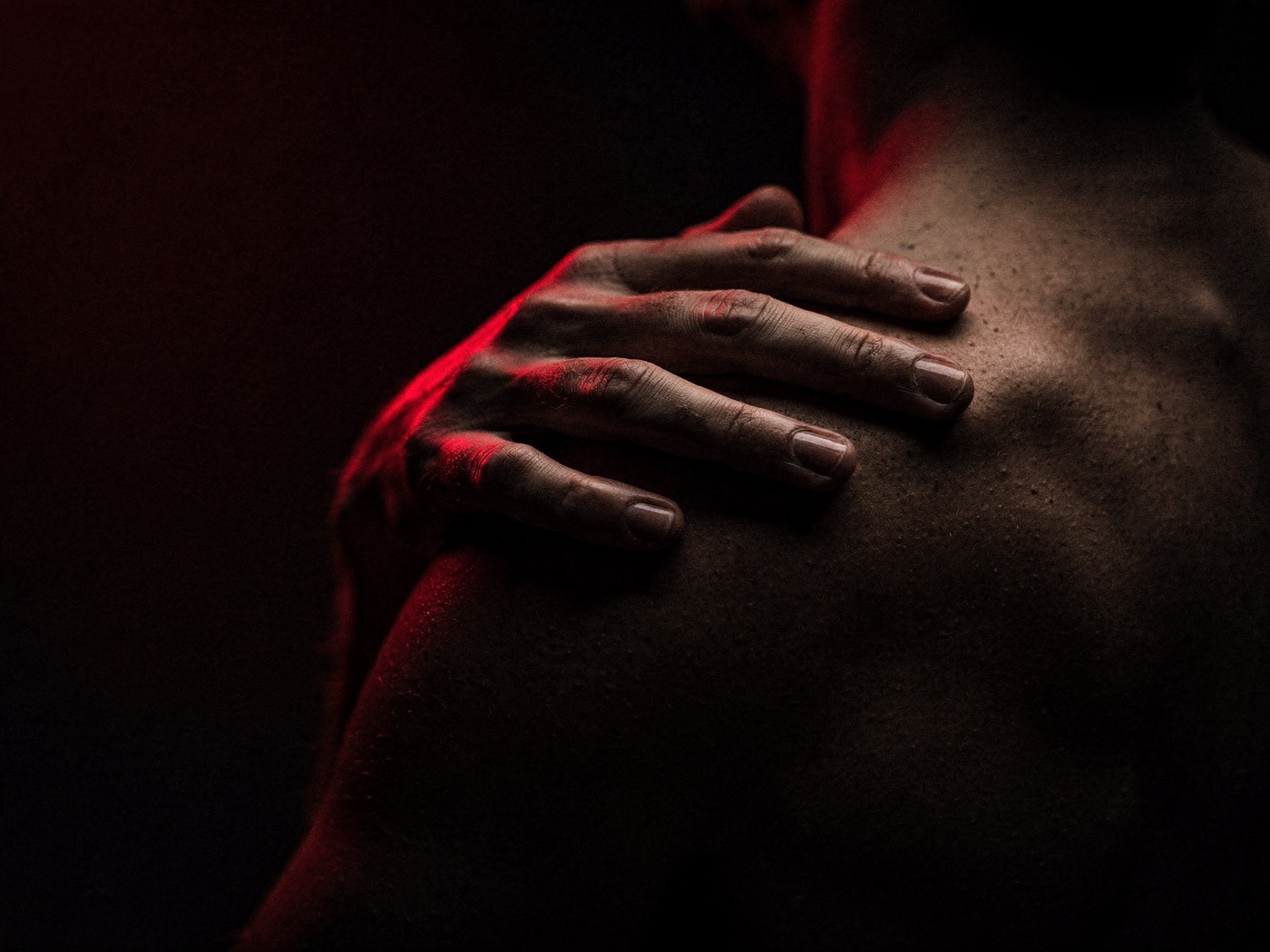 A hand resting on a bare shoulder in near darkness, lit deep red